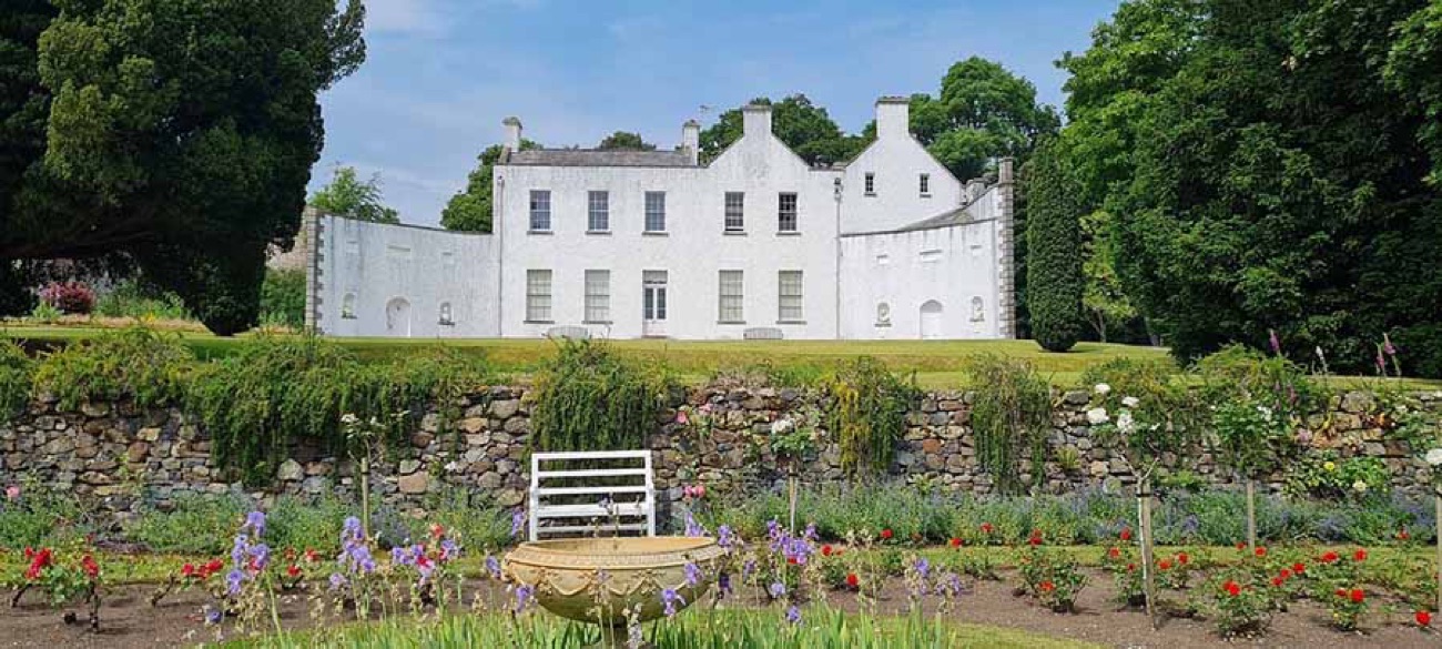 The white house at Ardress seen across the walled garden, with flower borders and a stone wall in the foreground