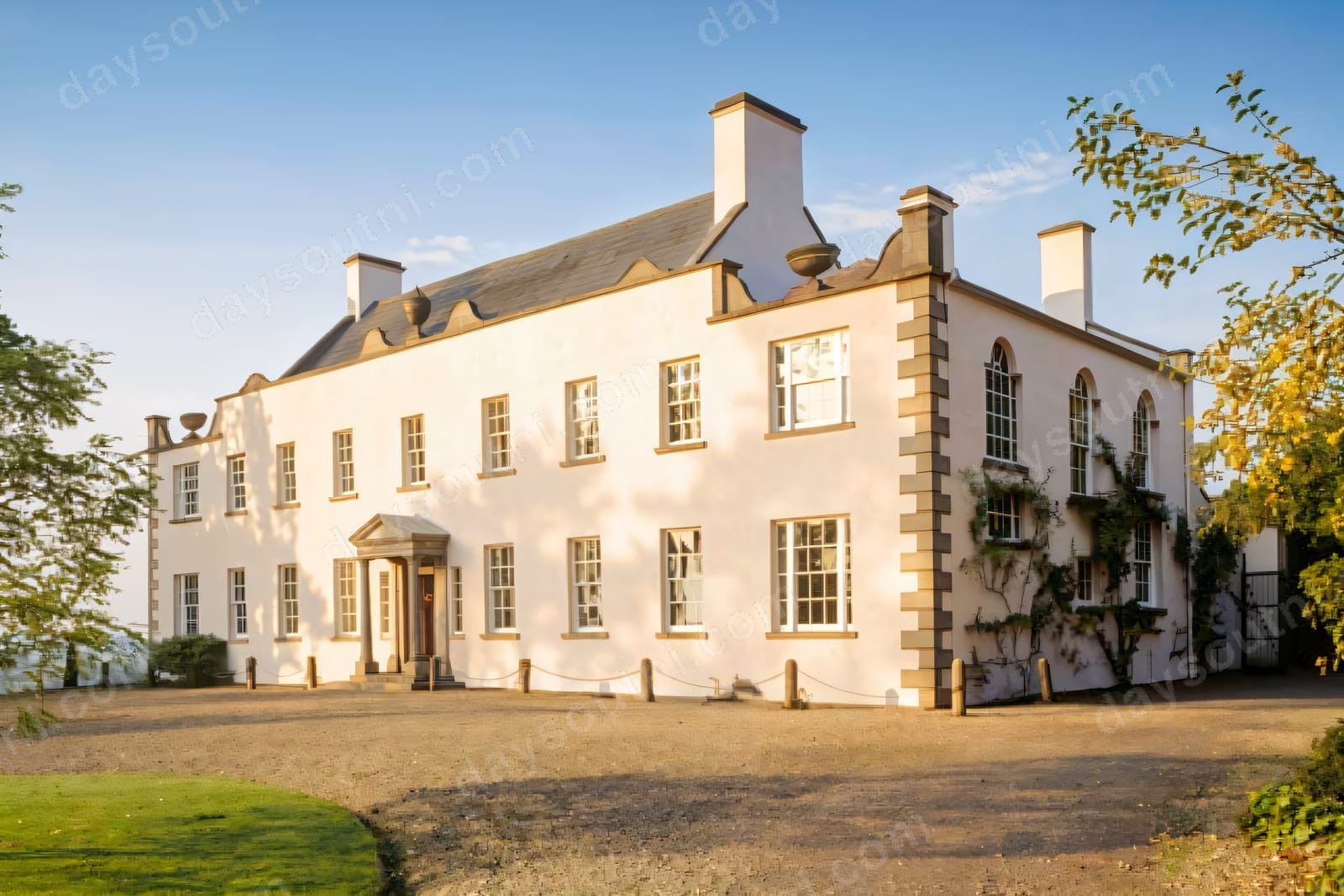 The whitewashed Georgian front of Ardress House with tall chimneys and a gravel forecourt, lit by low golden sunlight