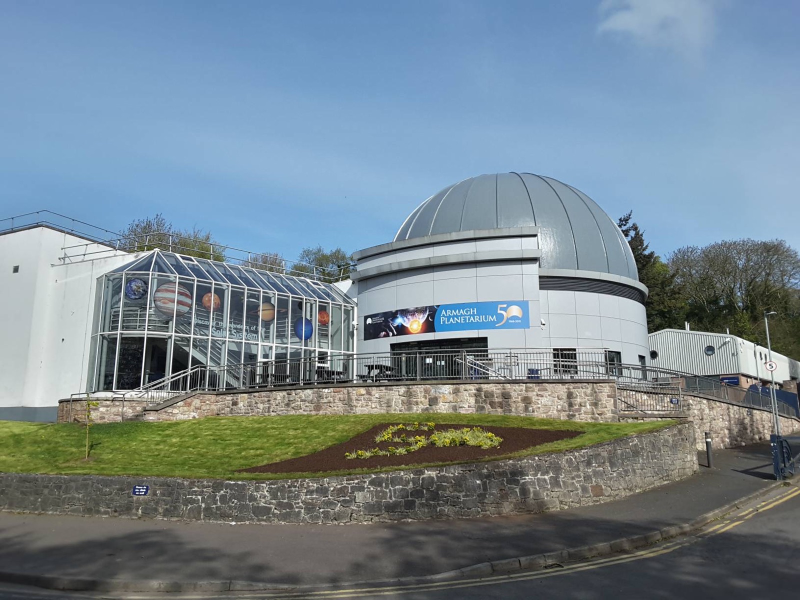 The silver planetarium dome by day beside the glass Astropark model-planets walkway under a blue sky