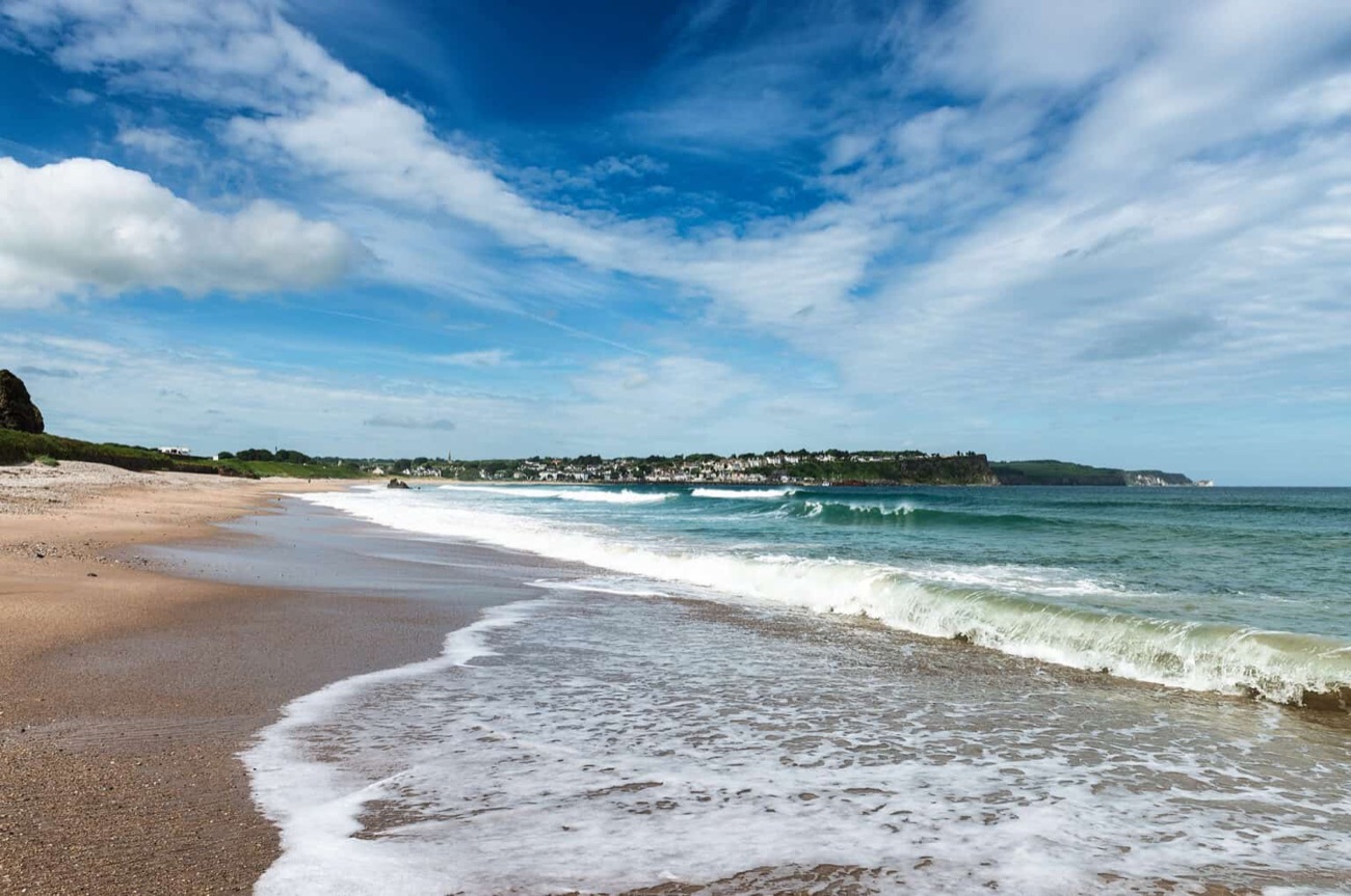 Waves rolling onto the wide sandy stretch of Ballycastle Beach with the town and headland in the distance under a big blue sky