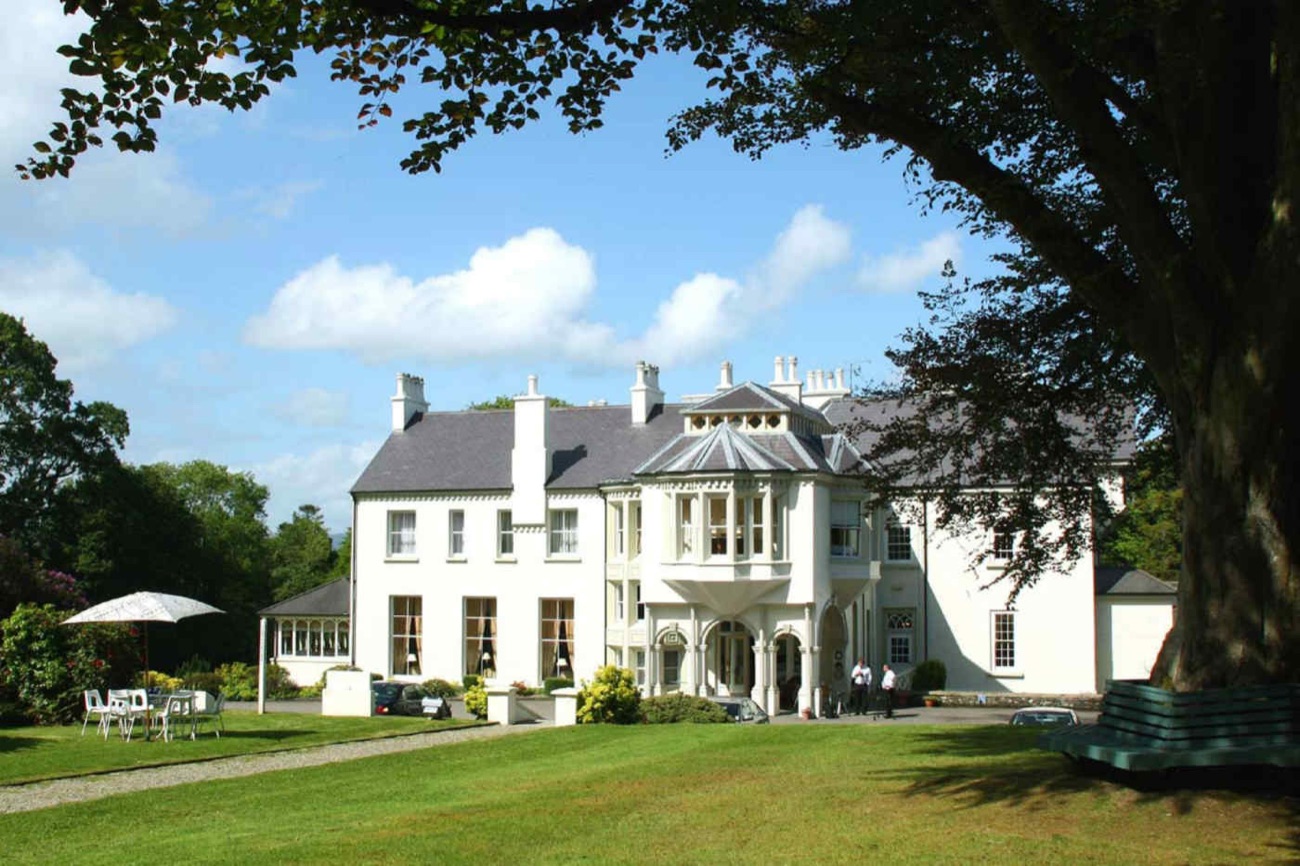 Beech Hill Country House seen across a wide green lawn from under a big beech tree, with a parasol and garden seating