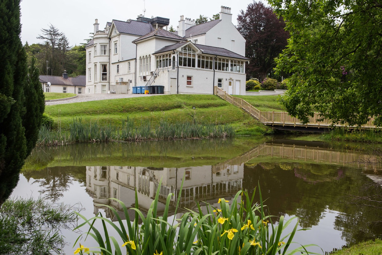 The house reflected in one of the grounds' ponds, with yellow flag irises in the foreground and a wooden footbridge