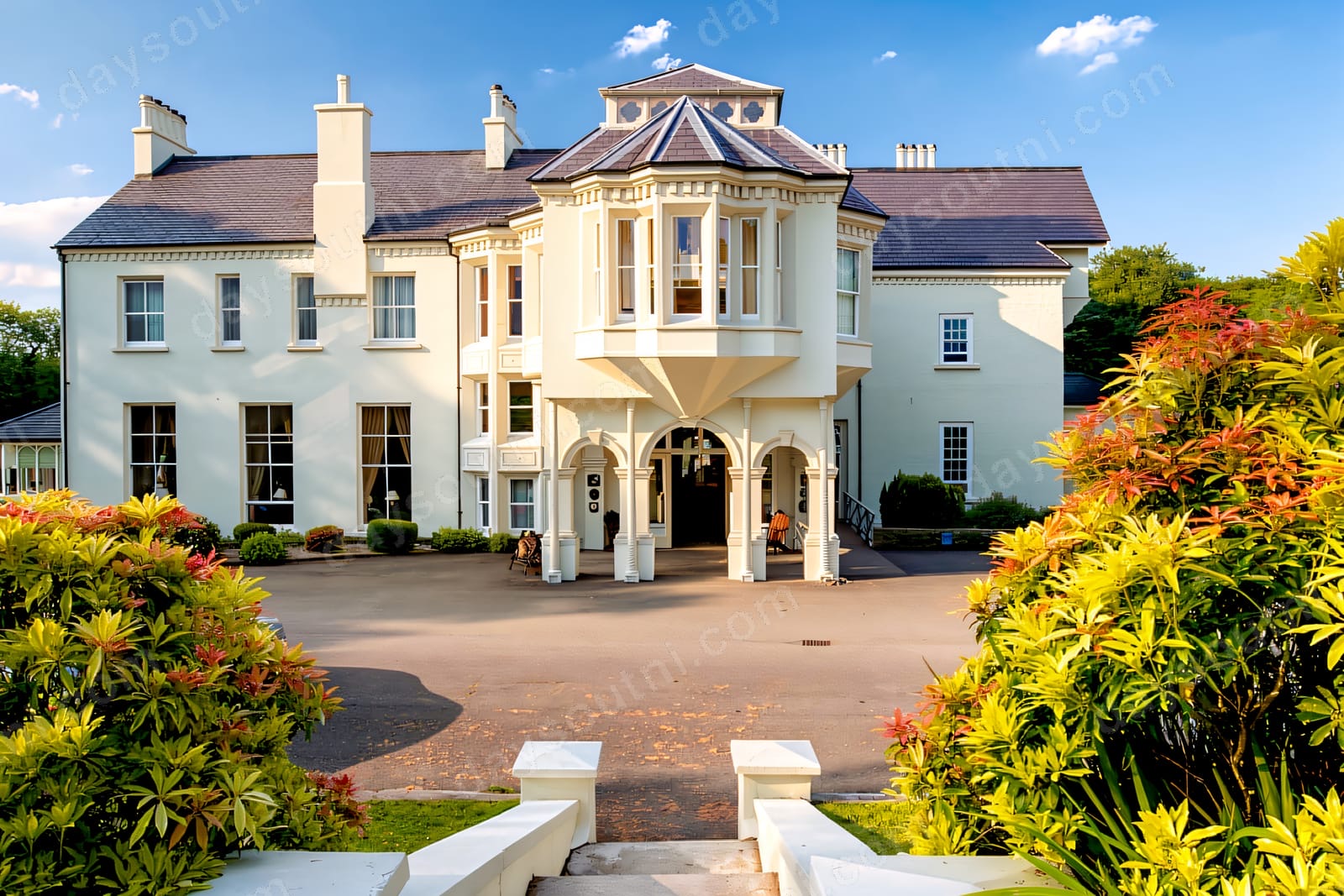 The cream-rendered Georgian front of Beech Hill Country House with its arched porch and bay tower, framed by red and green shrubs