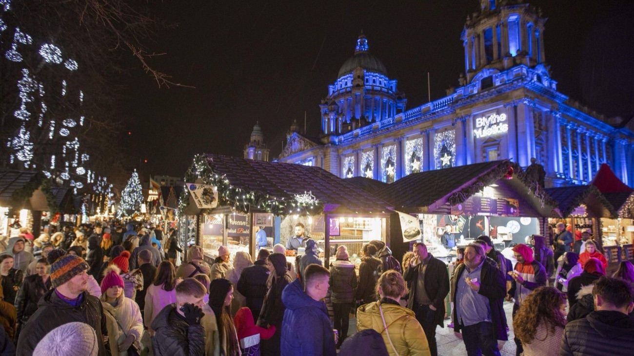 The floodlit dome of Belfast City Hall towering over the crowds and stalls of the Christmas Market at night