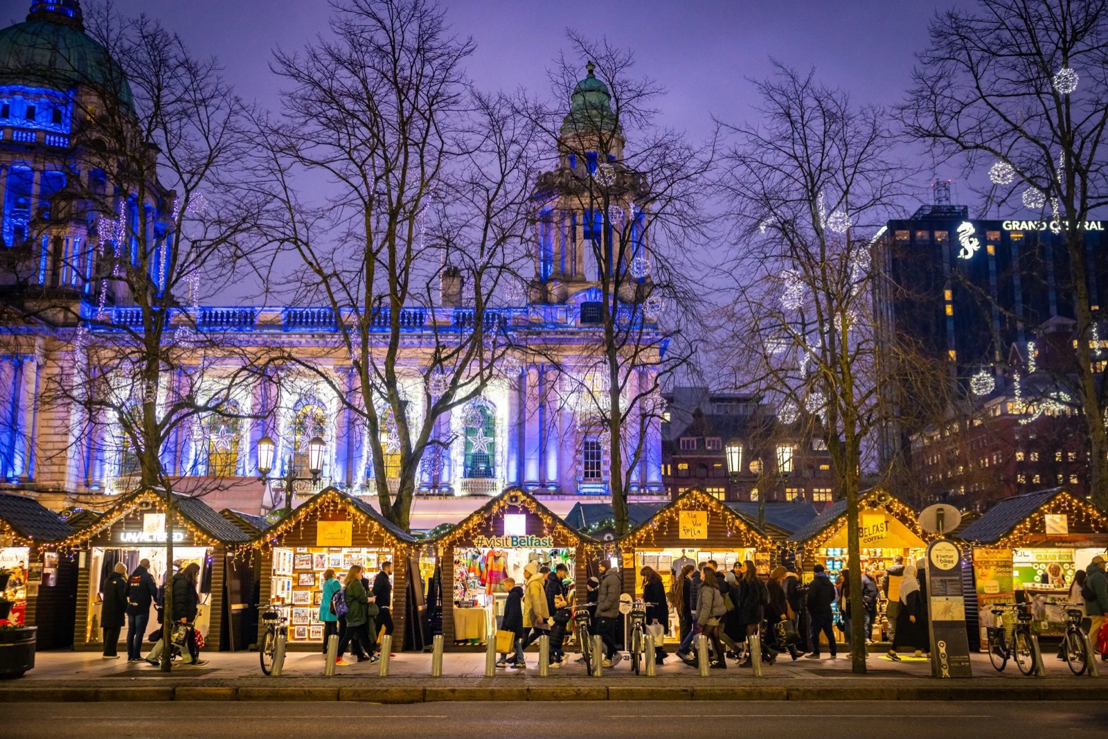 Rows of lit wooden chalet stalls at the Belfast Christmas Market with the blue-lit City Hall behind bare winter trees