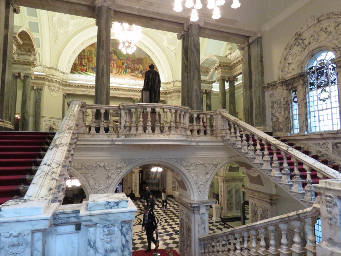 The grand marble staircase inside Belfast City Hall, with painted murals and a black-and-white tiled floor below