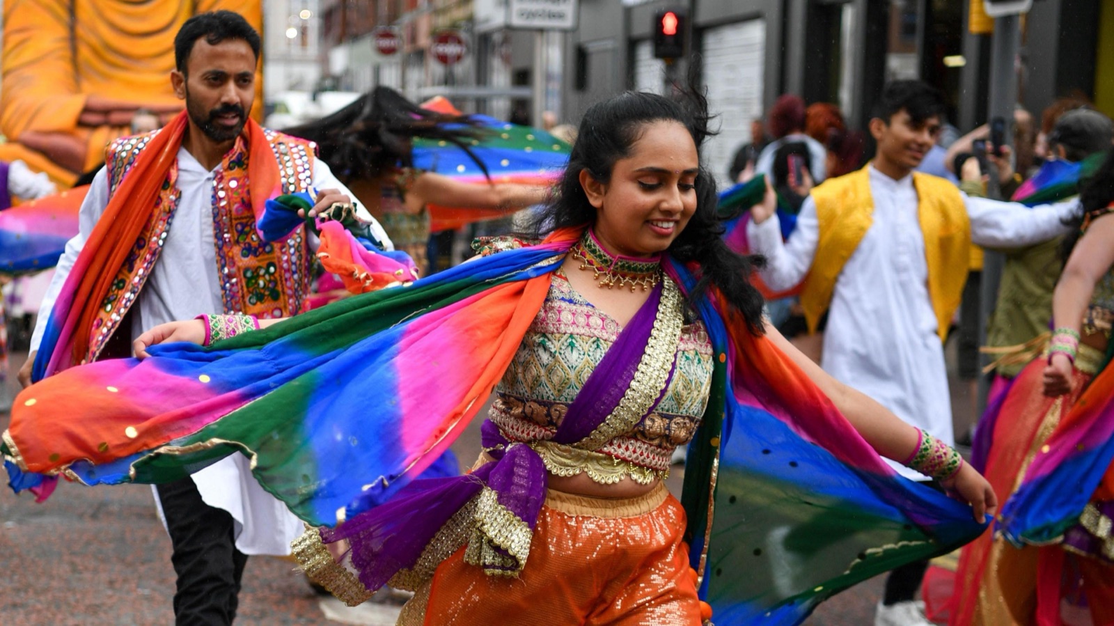 Bhangra dancers in gold and orange costumes with rainbow scarves flowing at Belfast Mela