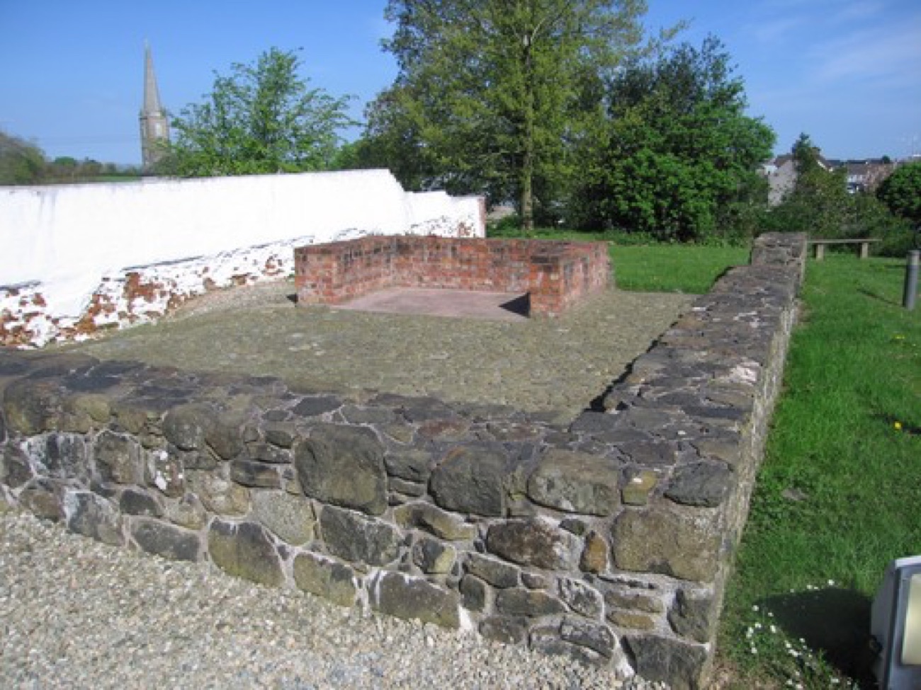 Excavated stone wall footings of the Plantation bawn at Bellaghy, with the village church spire behind