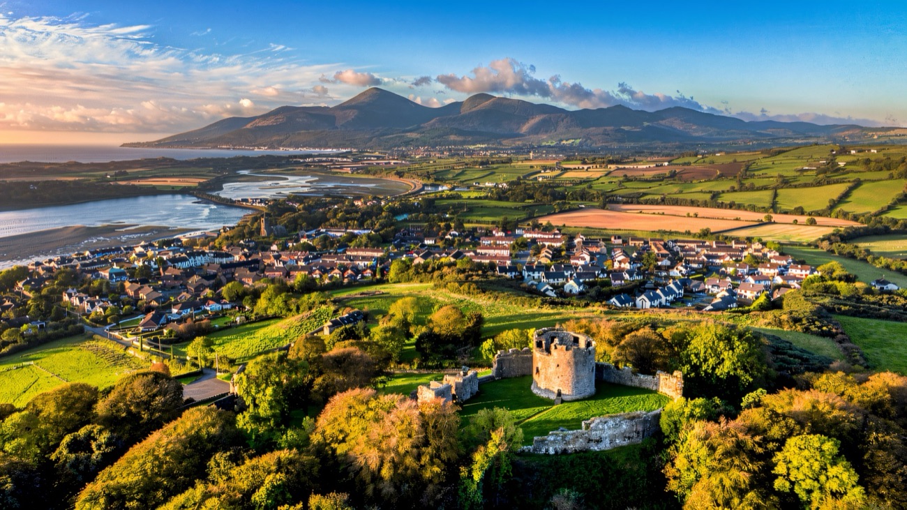 Aerial view of the round keep of Dundrum Castle on its wooded hilltop with Dundrum Bay and the Mourne Mountains beyond