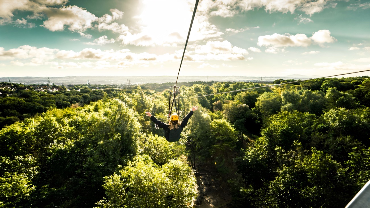A rider on a zip line flying over the green treetops of Colin Glen with Belfast beyond
