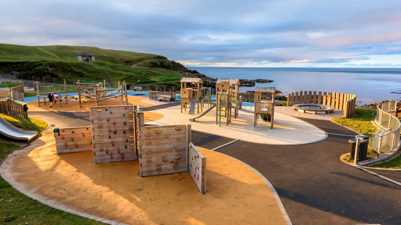Climbing walls and timber play structures on orange surfacing at the Portrush Recreation Grounds, with the sea and headland behind