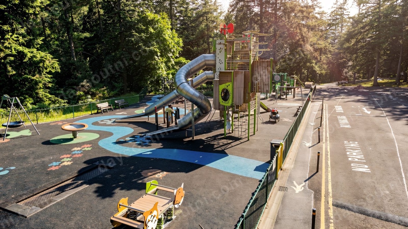 A tall green and timber climbing tower with a long steel tunnel slide at the Gortin Glen destination play park, conifers behind