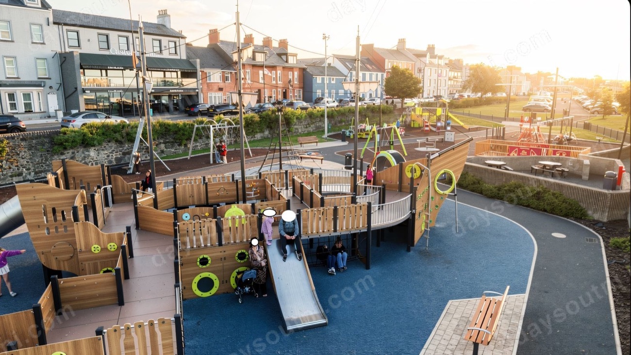 A large wooden ship play structure with slides and climbing nets on the Carrickfergus seafront, town houses behind