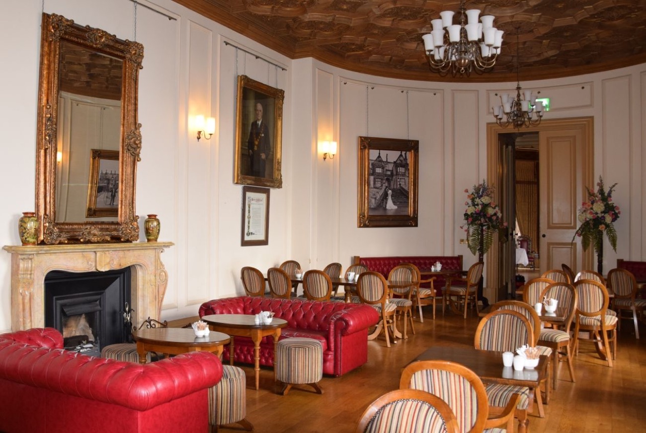 A grand interior room in Brownlow House with red leather sofas, a marble fireplace, a chandelier and an ornate carved ceiling