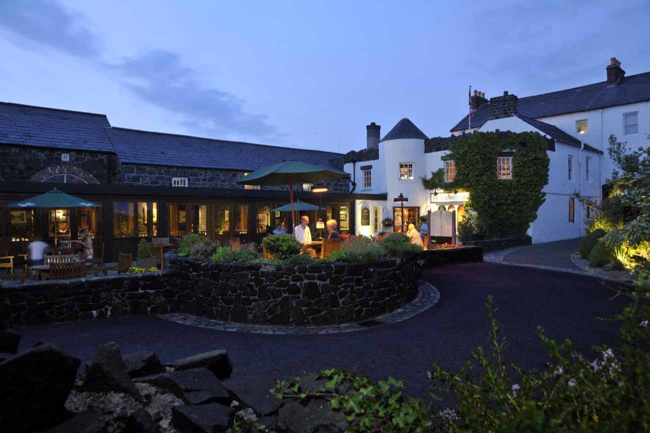 The Bushmills Inn at dusk, its windows glowing warm across the flagstone courtyard and the little round turret catching the last light