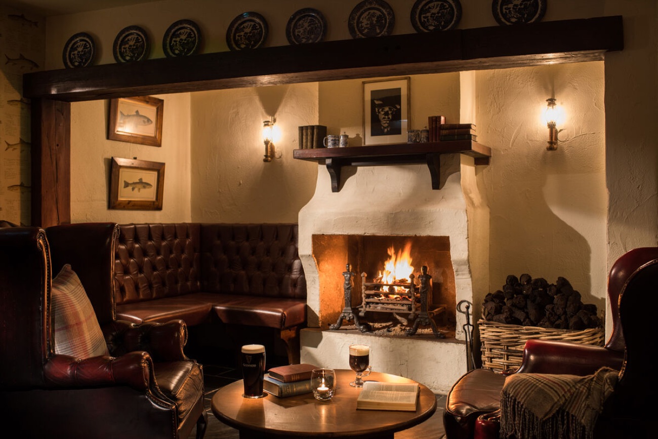 A cosy fireside snug at the Bushmills Inn with an open fire, a deep leather bench, two pints and a stack of old books on the round table