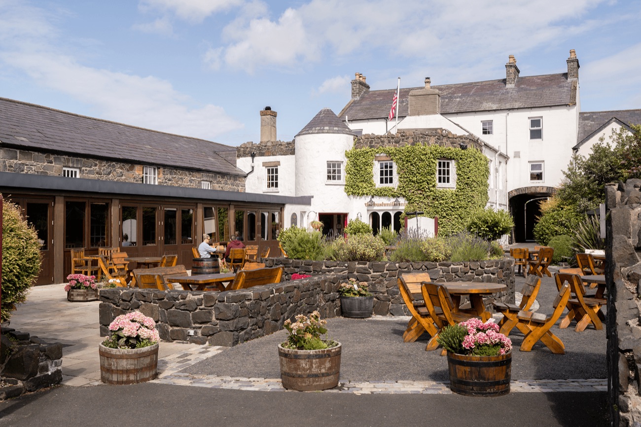 The sunny courtyard garden at the Bushmills Inn, with wooden tables, whiskey-barrel planters of pink flowers and the ivy-covered inn behind