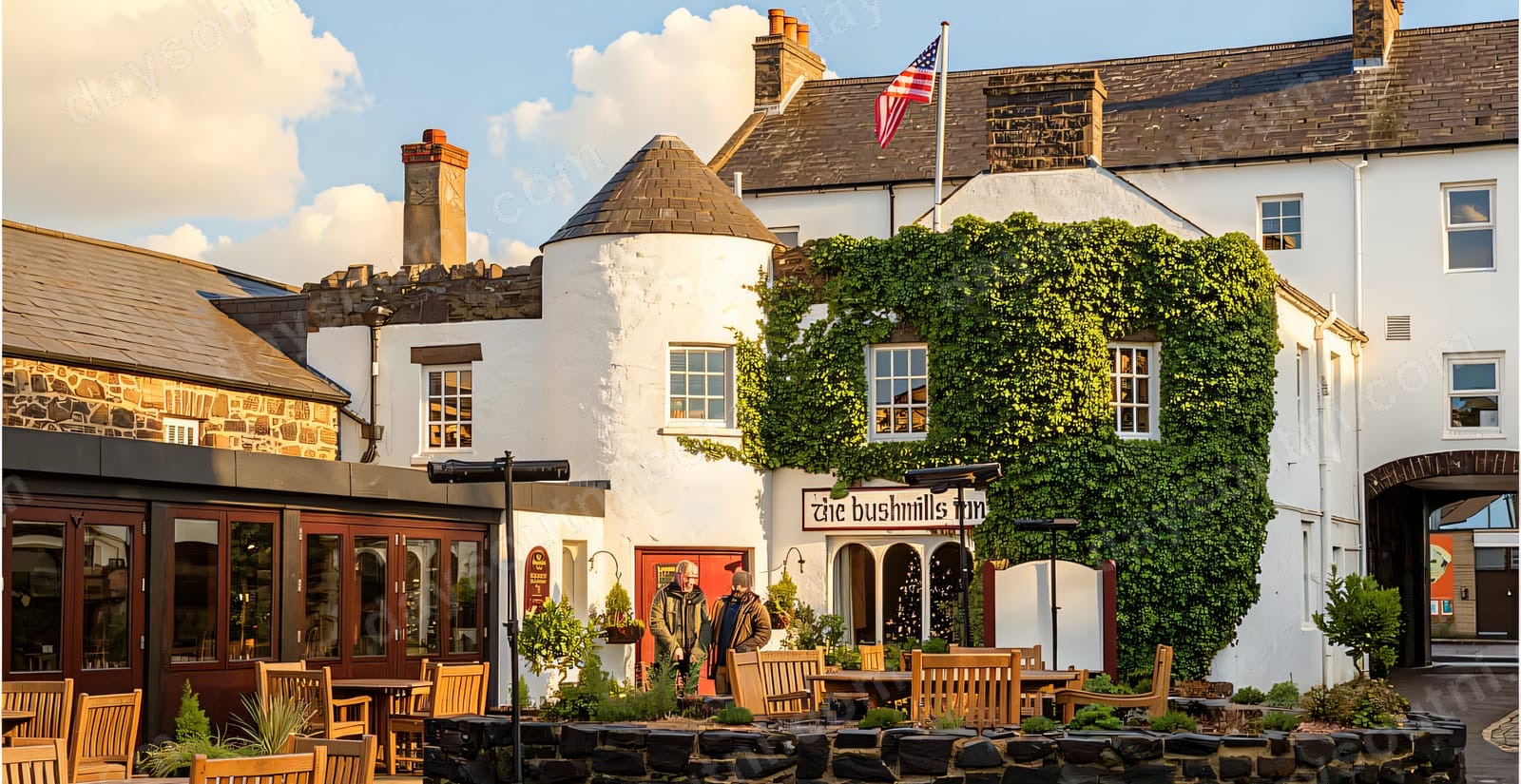 The whitewashed frontage of the Bushmills Inn with its ivy-clad round turret, the inn's sign lit above the door and a courtyard of wooden tables in front