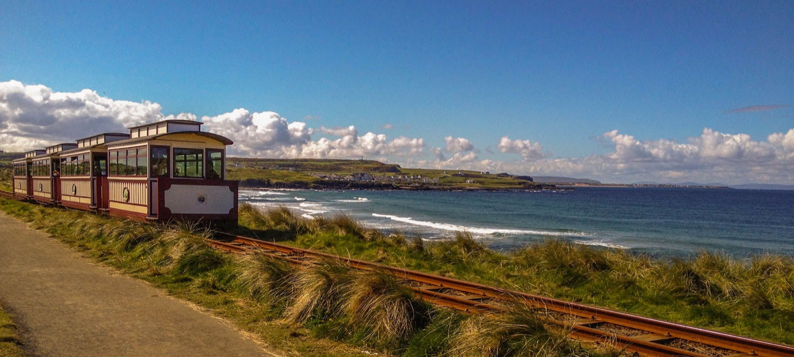 A heritage tram of red-and-cream carriages running on the narrow-gauge track along the Antrim coast, sea and headland behind