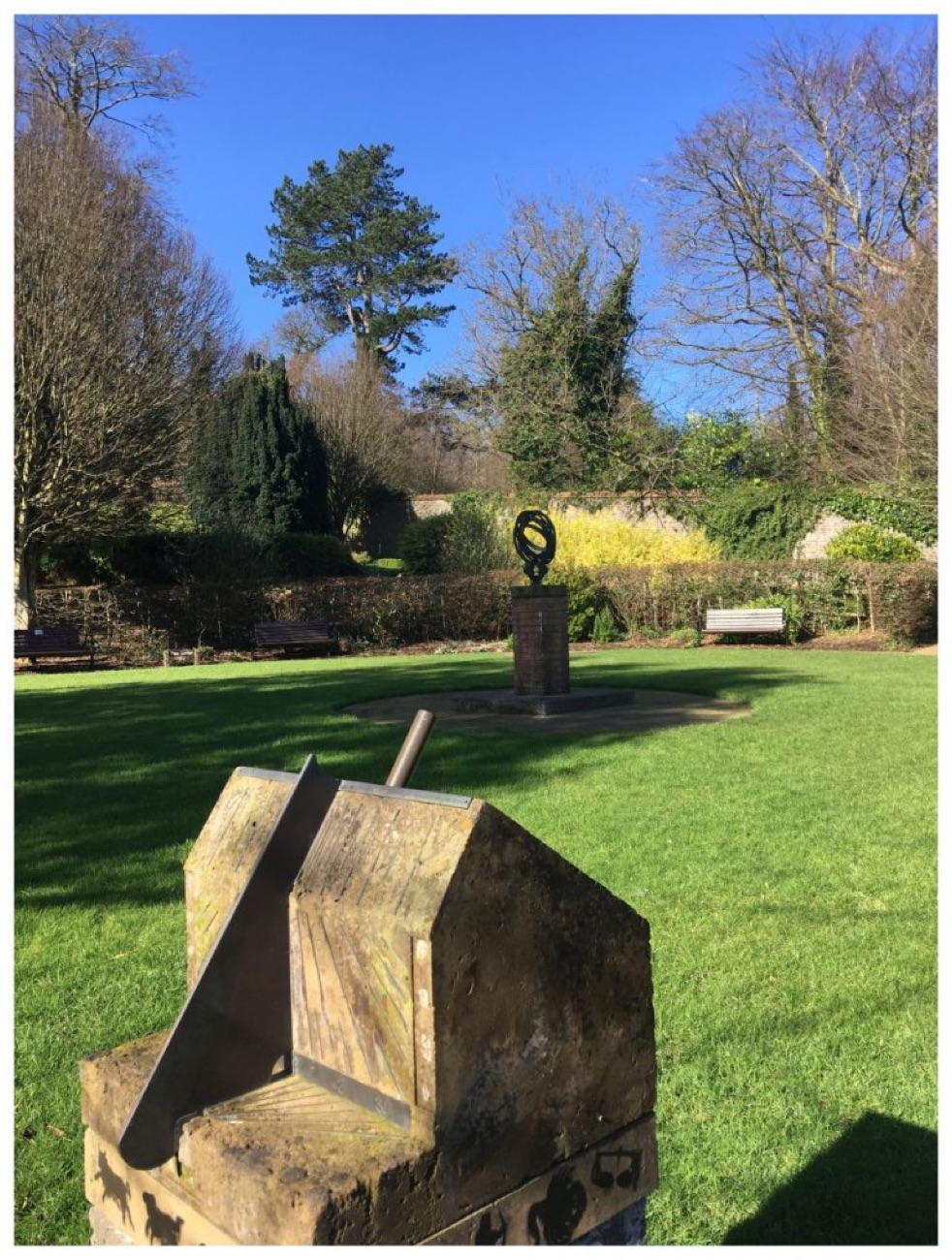 A stone sundial in the foreground of Carnfunnock's walled garden, an armillary sundial on a plinth on the lawn and benches under the trees