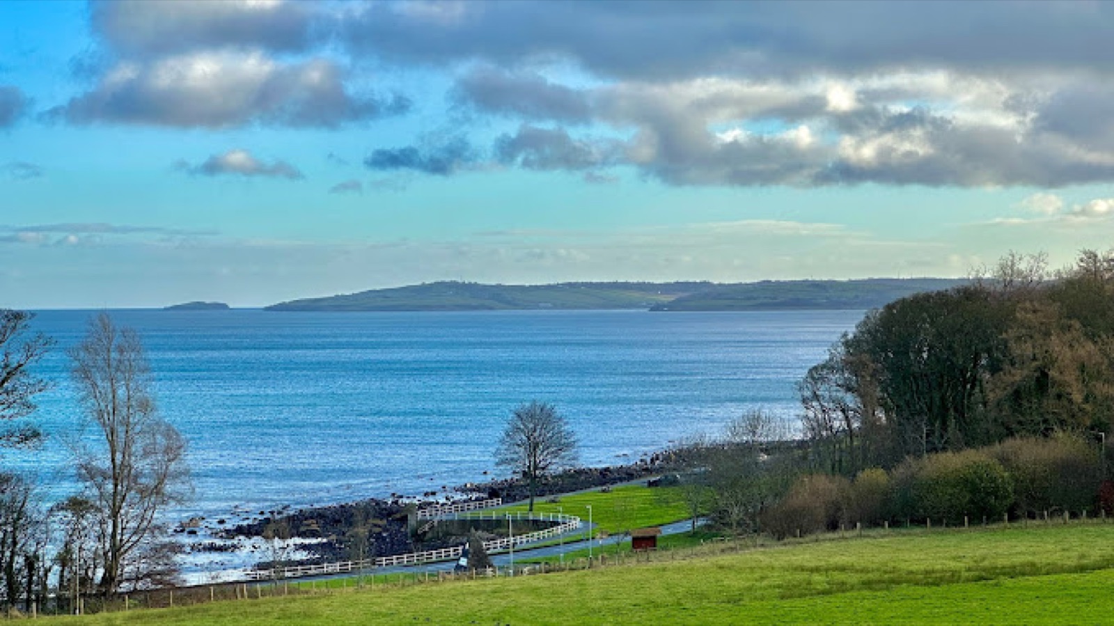 The sea and a green headland seen from Carnfunnock Country Park, with grass, a shore path and trees running down to the water