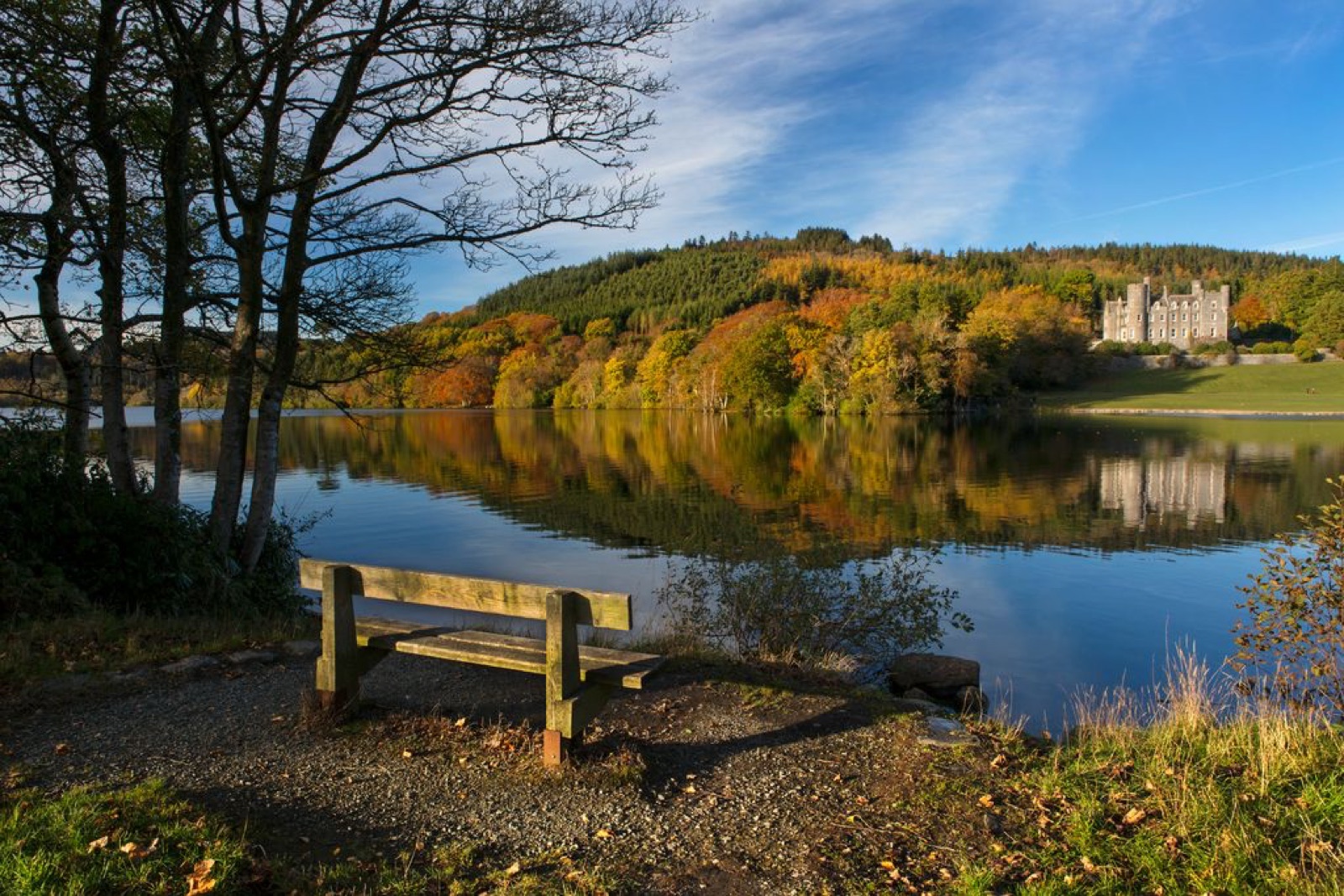 Castlewellan Lake in autumn colour with the grey baronial castle across the water, a lakeside bench and the Mourne Mountains beyond