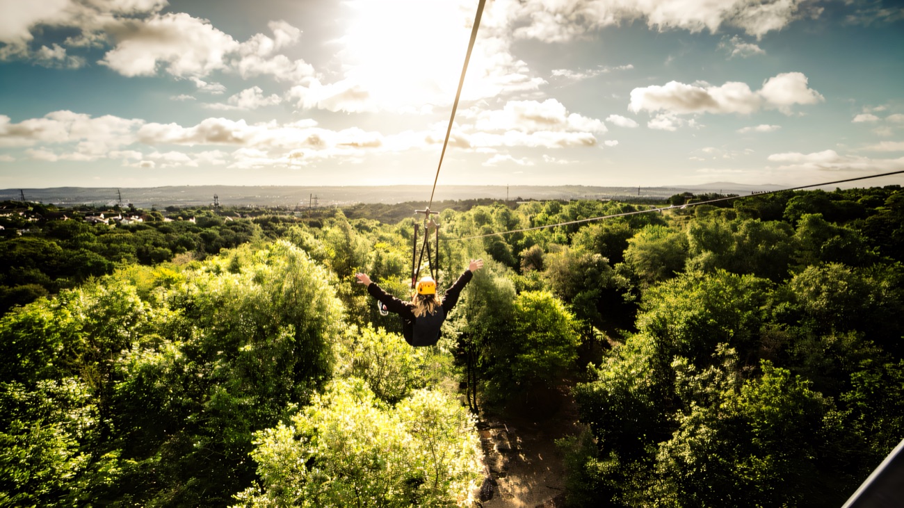 A rider in a helmet flying out along the zip line high over green woodland toward the Belfast hills