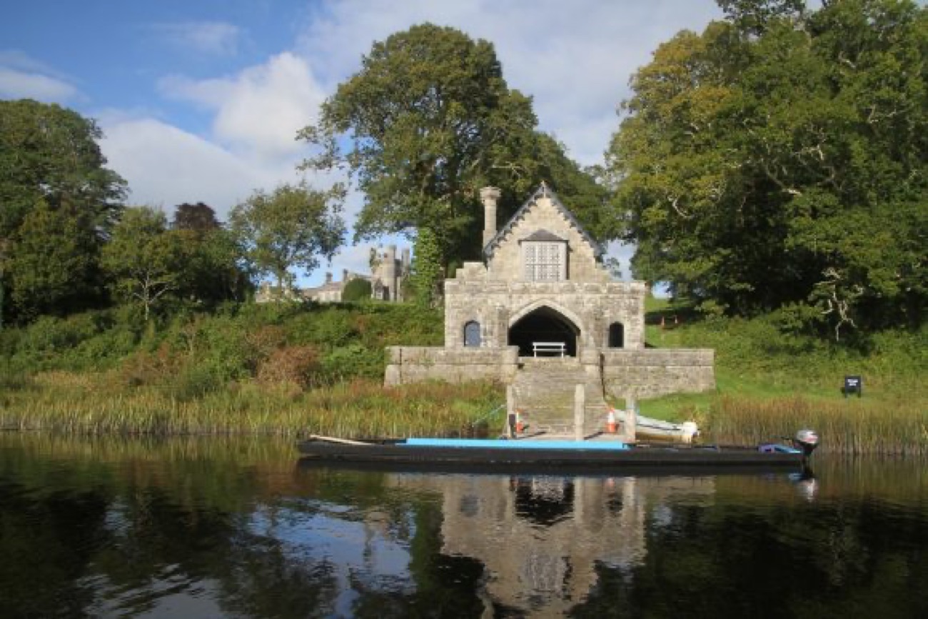 The stone Gothic boathouse at Crom Estate reflected in the lough, with a long wooden boat moored in front