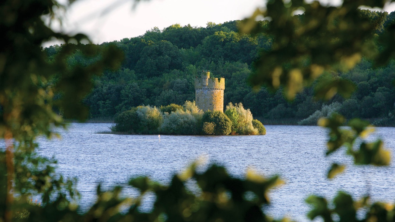 A round stone tower on a small wooded island out in Upper Lough Erne, glowing in the evening light at Crom
