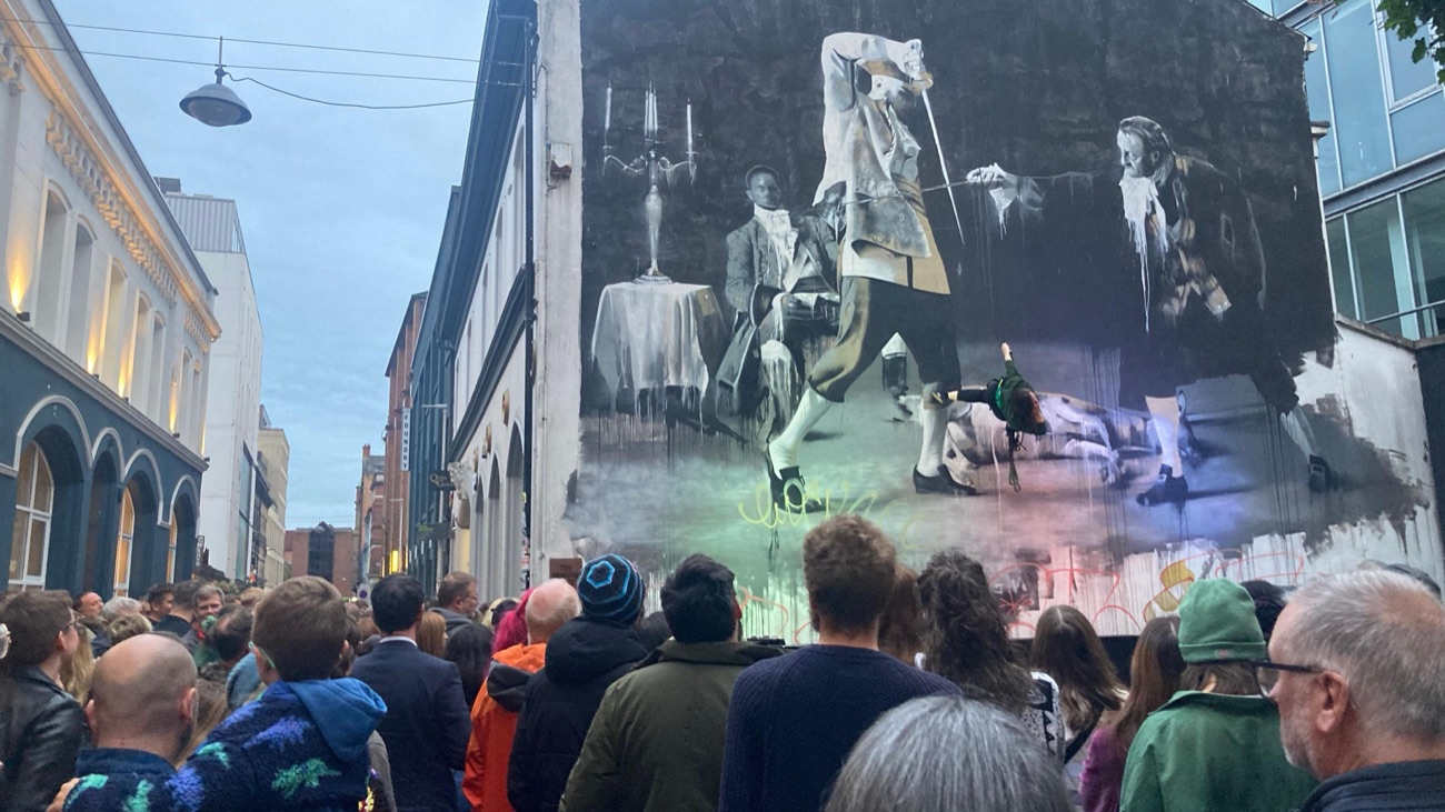 A crowd gathered in front of a large street mural at dusk in Belfast's Cathedral Quarter on Culture Night