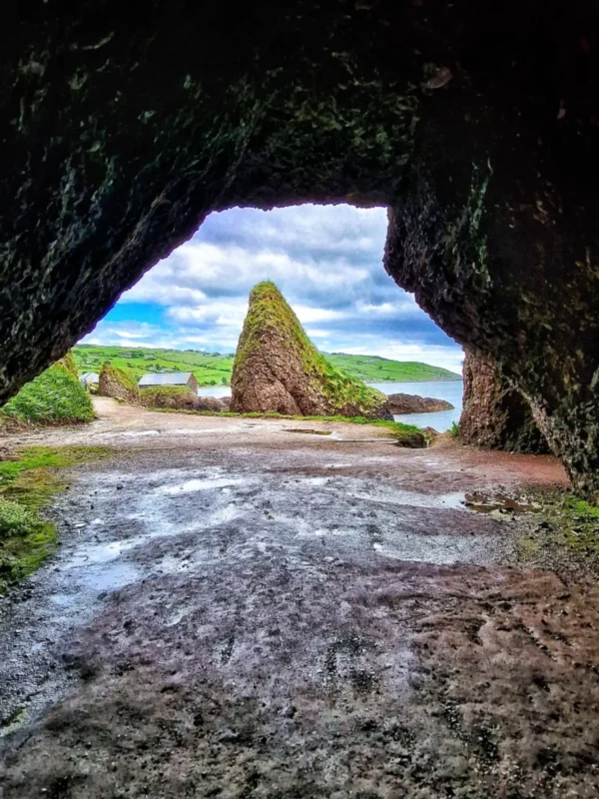 A cave arch framing a sea stack, green hills and a whitewashed cottage at Cushendun
