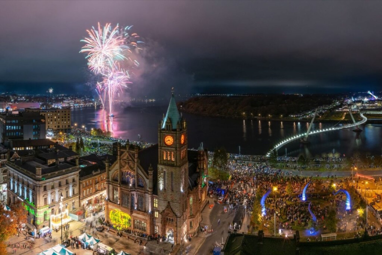 Aerial view of the floodlit Guildhall and huge crowds along the Foyle with fireworks and the Peace Bridge lit up
