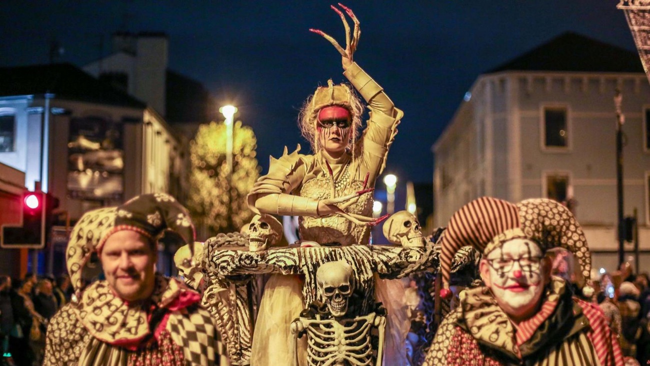 Carnival performers in elaborate skeleton-queen and harlequin costumes on a Derry street at night