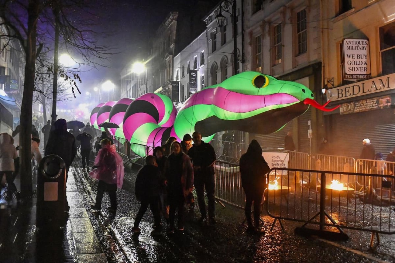 A giant illuminated pink-and-green serpent light installation down Shipquay Street with families walking the Awakening the Walled City trail