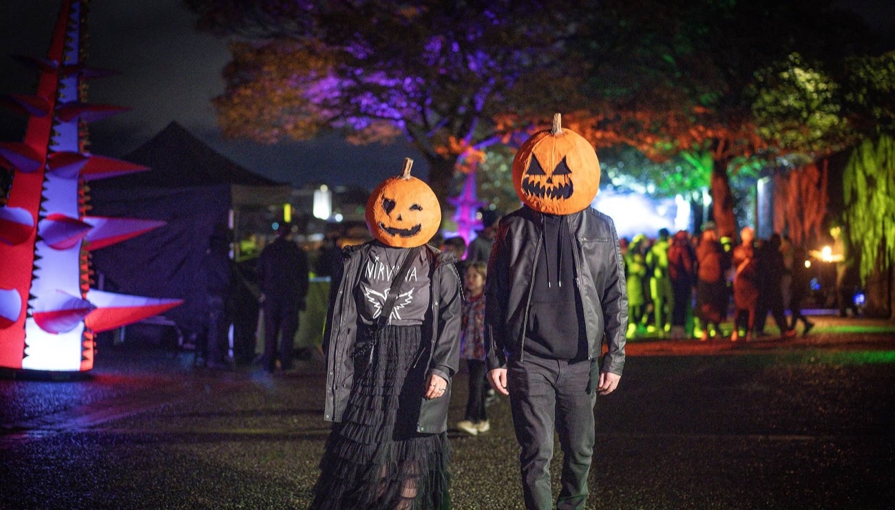 Two visitors with glowing carved-pumpkin heads walking the illuminated Halloween trail in Derry at night