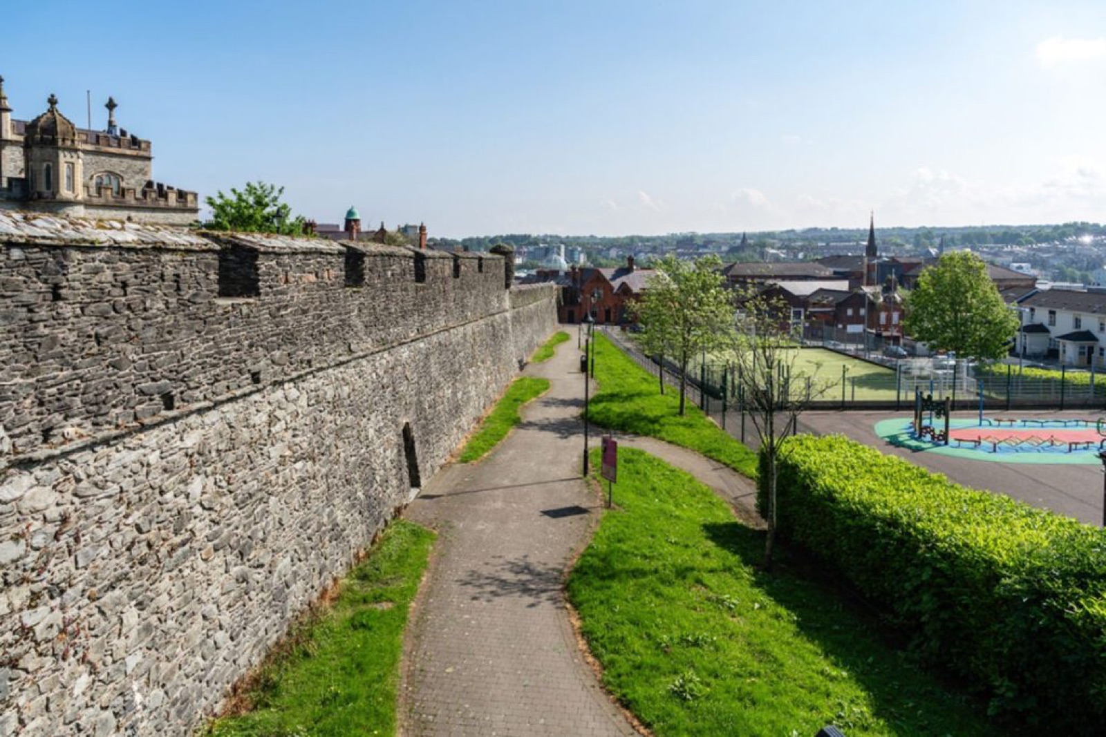 The broad stone wall walkway with a path, green space and the city beyond under blue sky