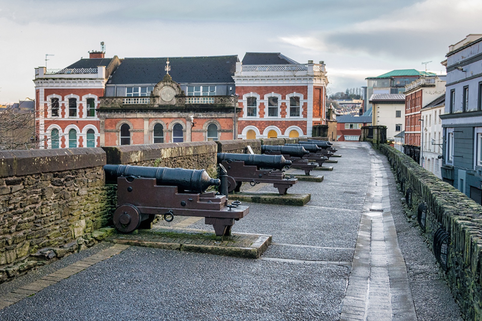 A row of old black cannons along the stone rampart walkway, with the Guildhall-quarter buildings behind