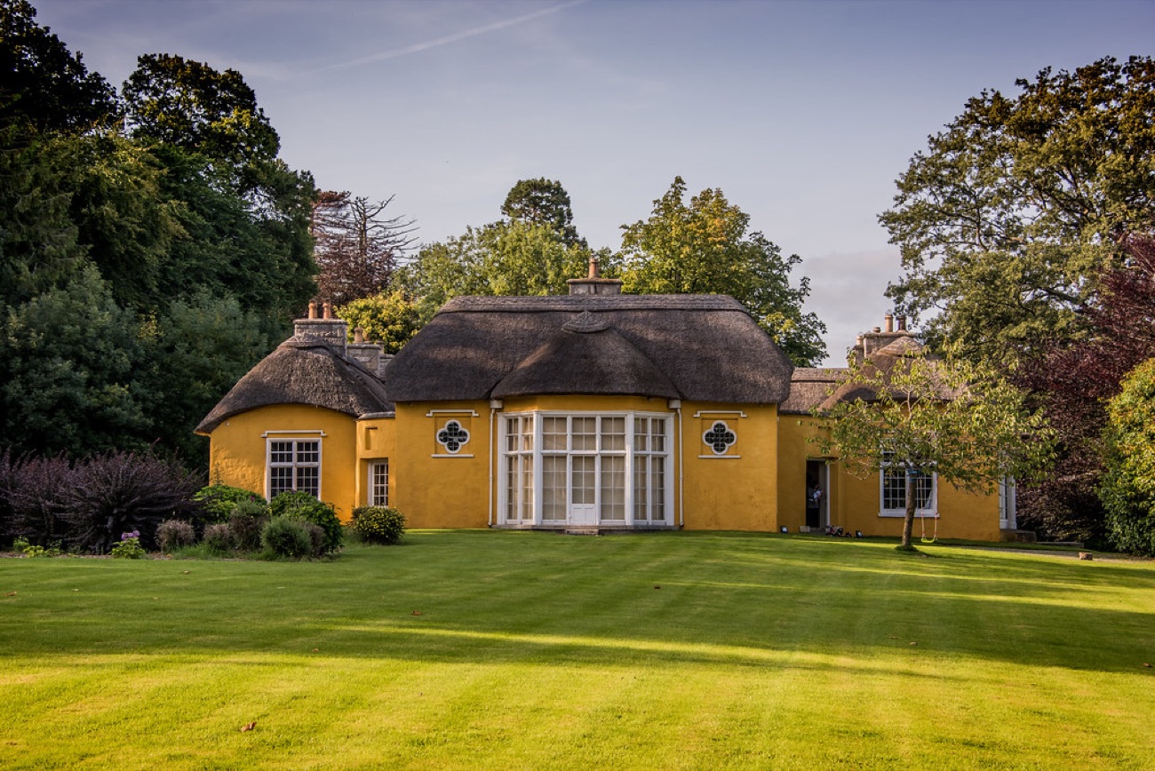 Derrymore House lit by golden evening light, the central bow window and thatched roof framed by tall trees and open lawn