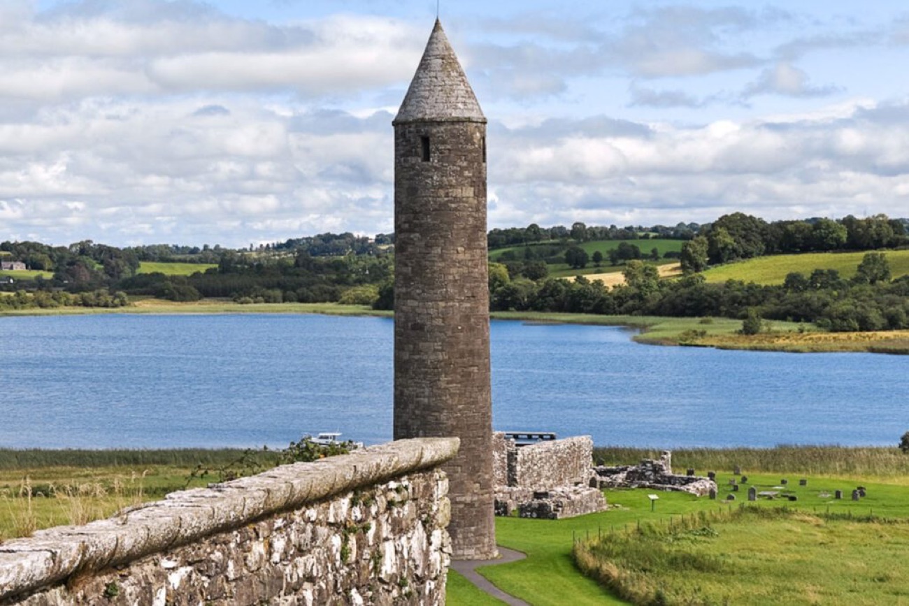 The round tower close up beside the priory ruins on Devenish Island, with Lough Erne behind under a blue sky