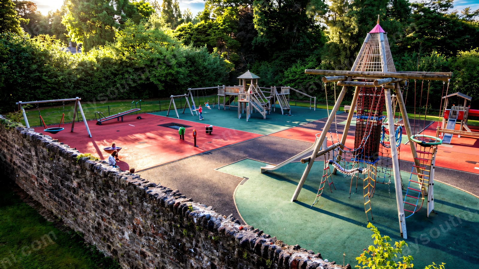 A tall rope-and-timber climbing tower on red and green safety surfacing inside an old walled garden.
