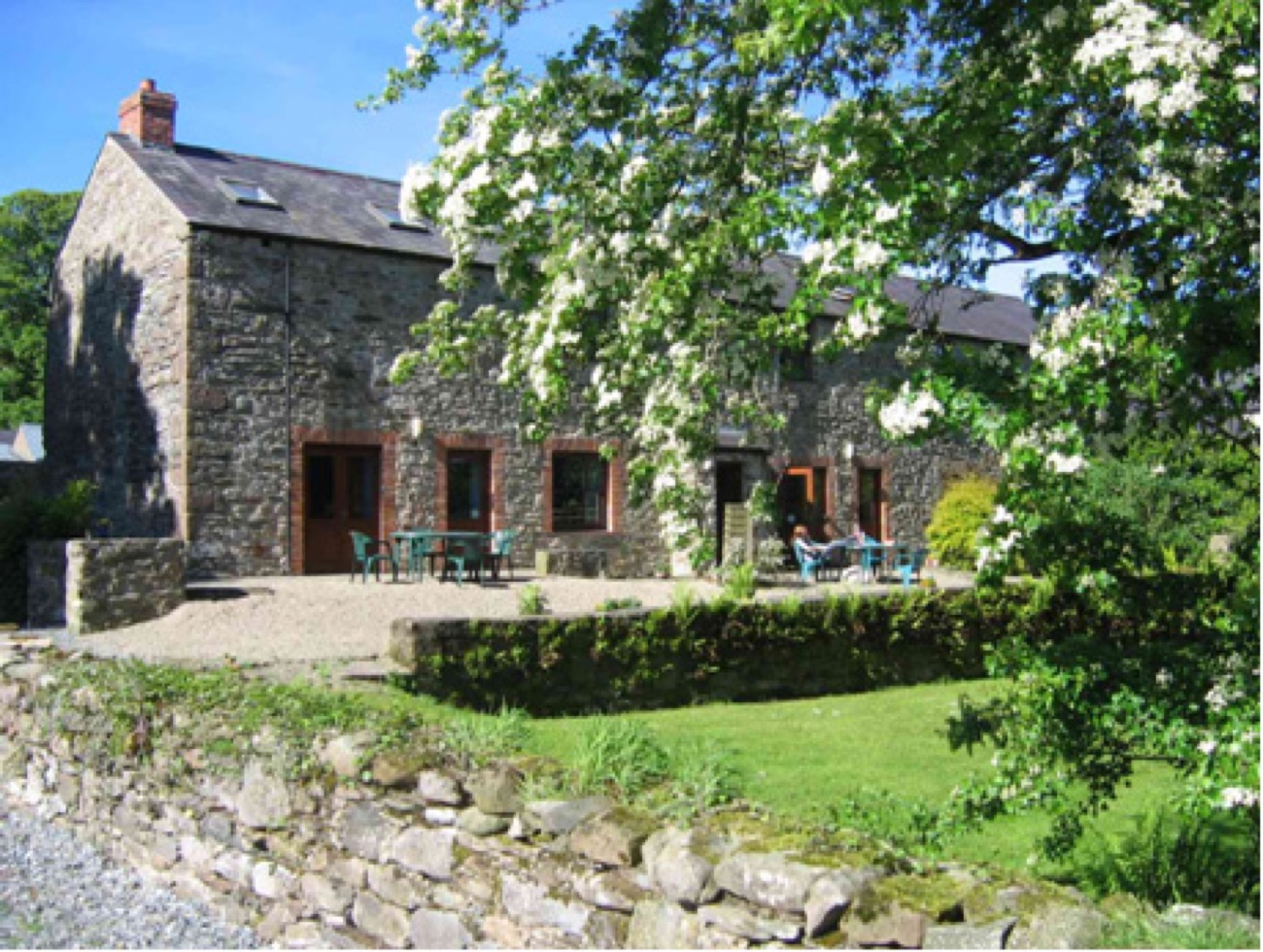 The converted stone barn at Drumcovitt with terracotta doors and patio tables, a flowering tree and dry-stone wall in front