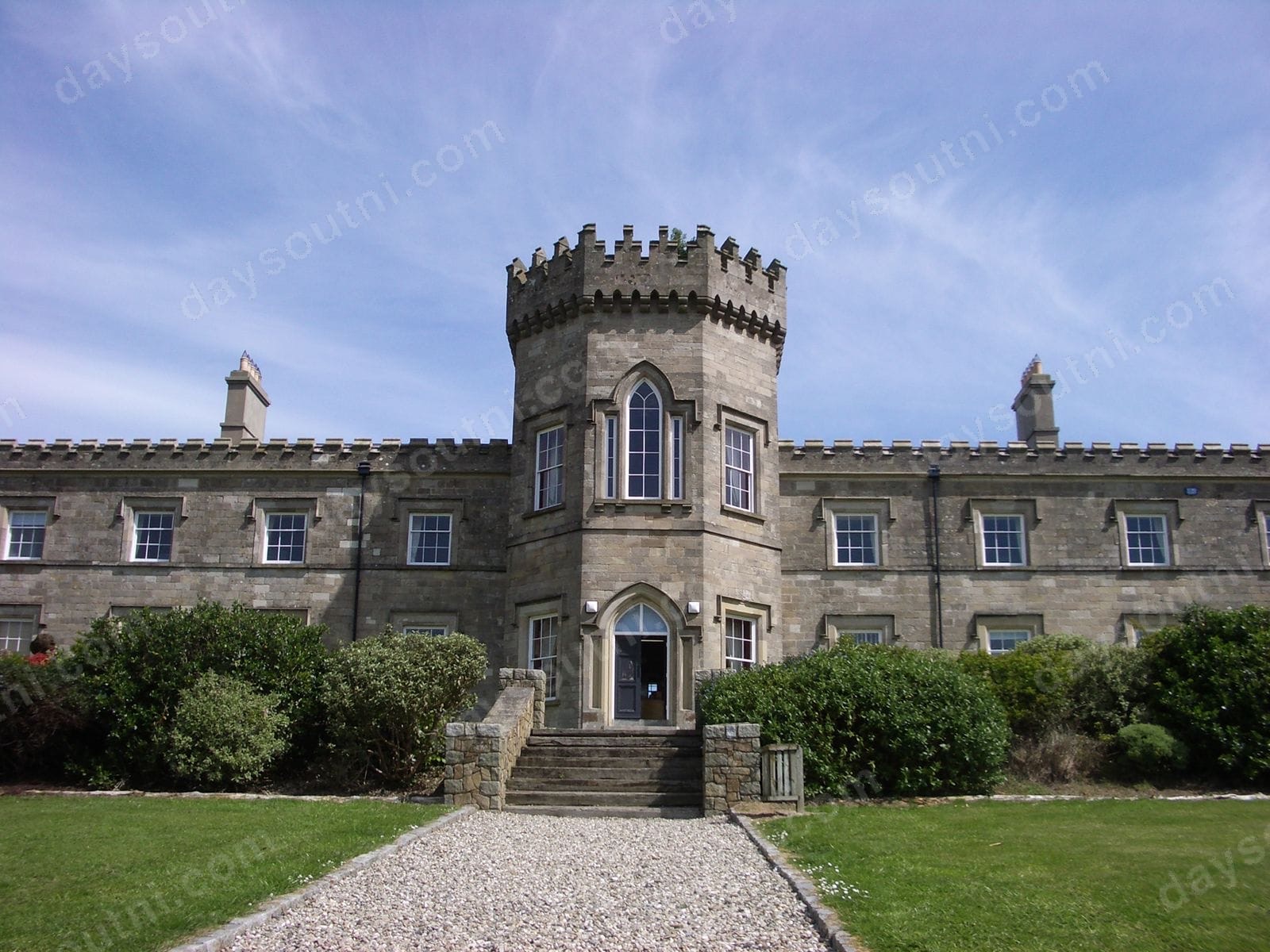 The battlemented central tower and grey-stone front of Dungiven Castle above a gravel path and lawn