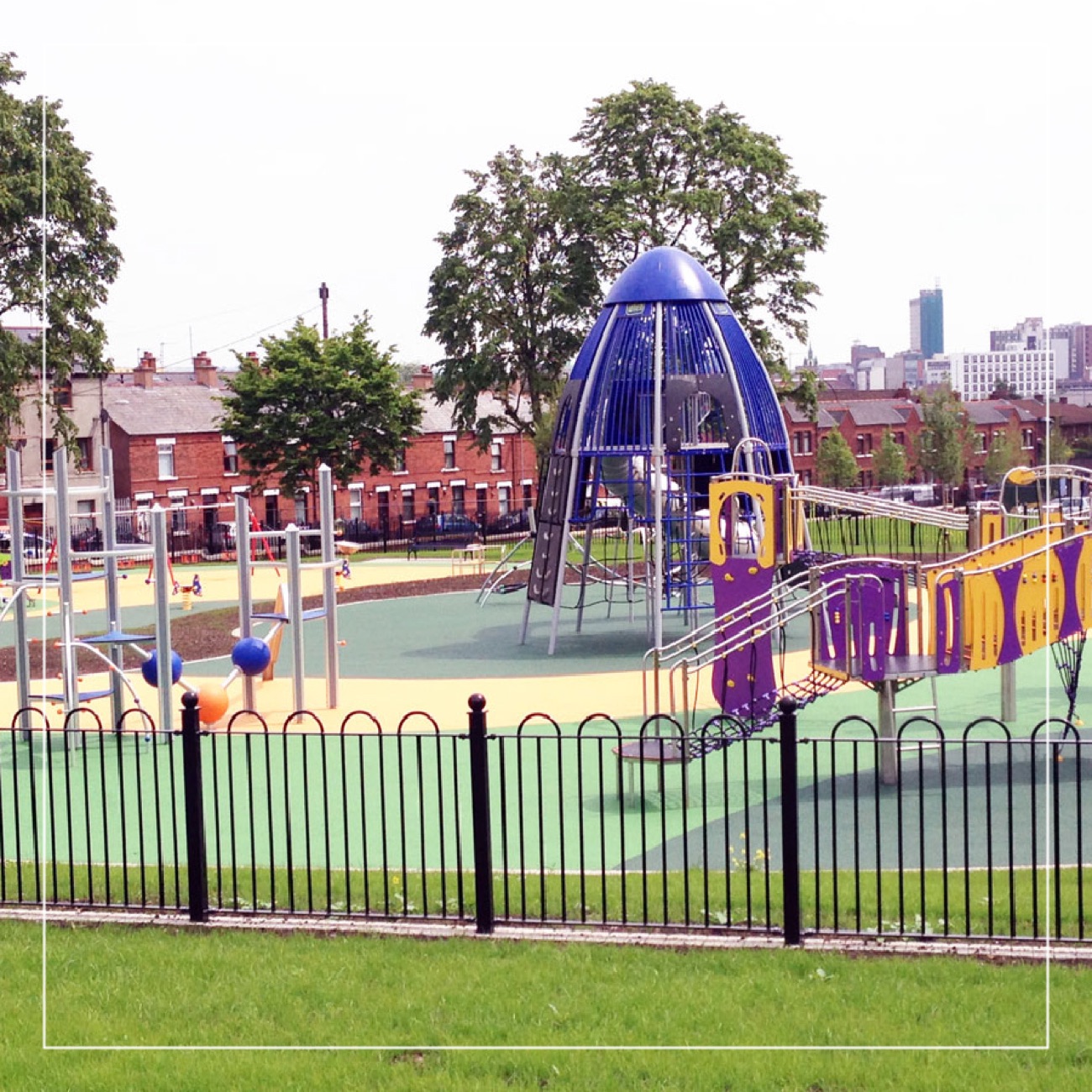 The play park at Dunville with a blue domed climbing structure and the red-brick Falls Road terraces behind