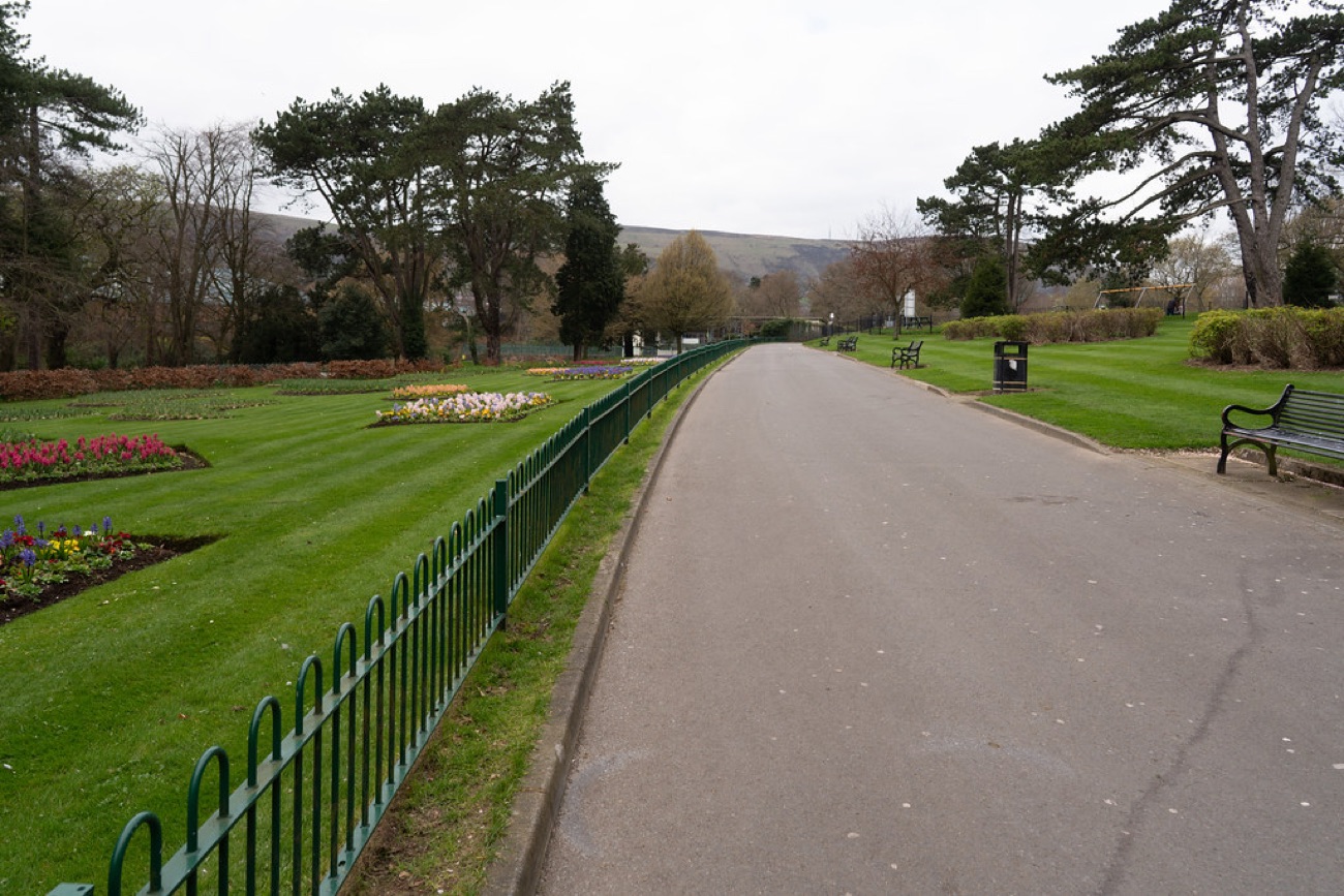 A wide tarmac avenue through Falls Park with flower beds, benches and the Belfast Hills behind