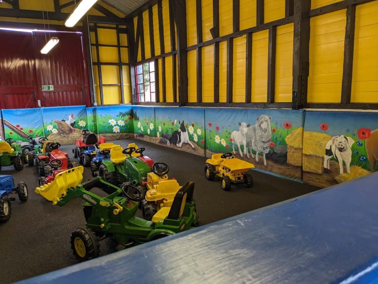 A fleet of ride-on pedal tractors in the indoor play barn with painted farm-animal murals along the walls