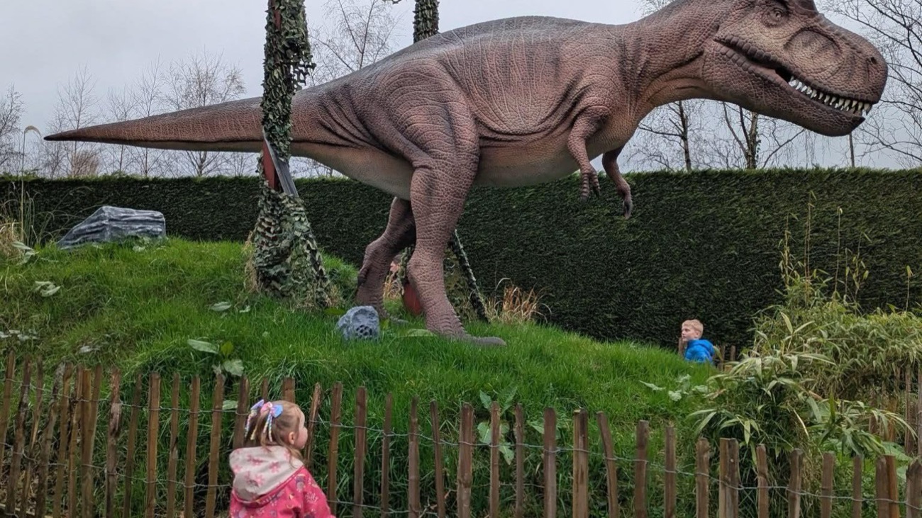 A large model dinosaur on the outdoor dinosaur trail with children looking on from behind a fence