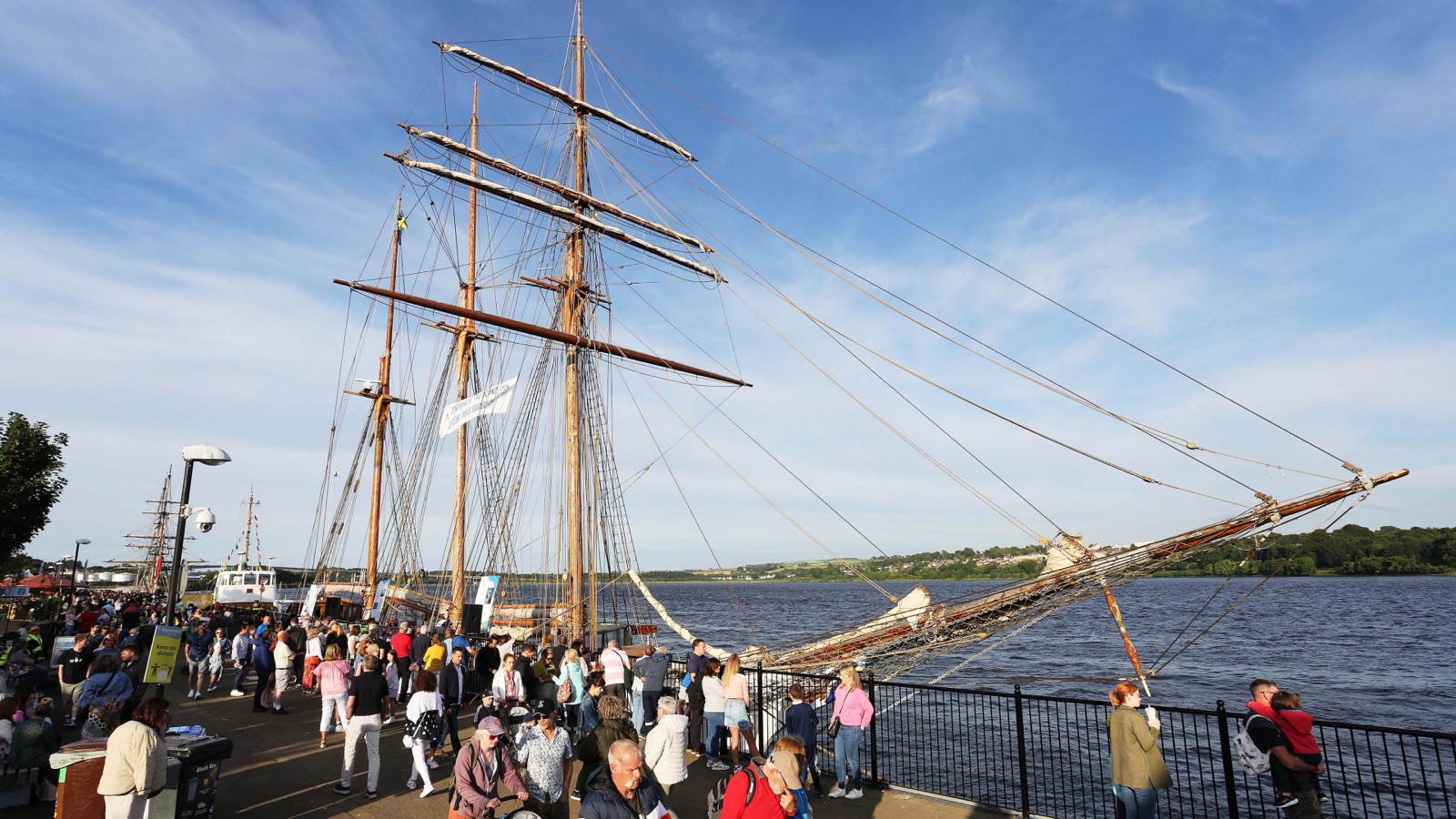 A tall ship moored at the Derry quay with crowds walking the riverside and the River Foyle beyond on a sunny day