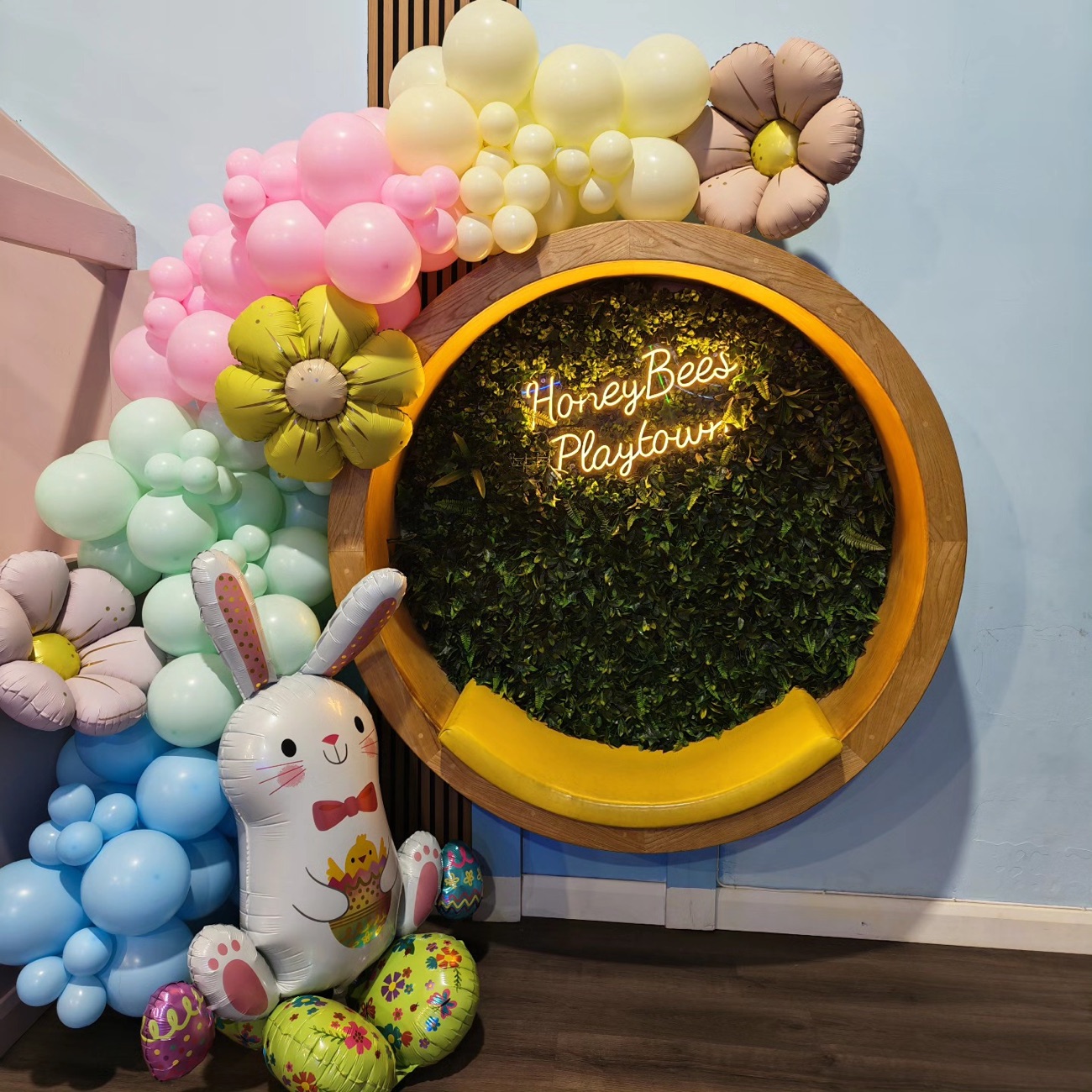 The 'HoneyBees Playtown' neon sign on a greenery feature wall, framed by a pastel balloon arch