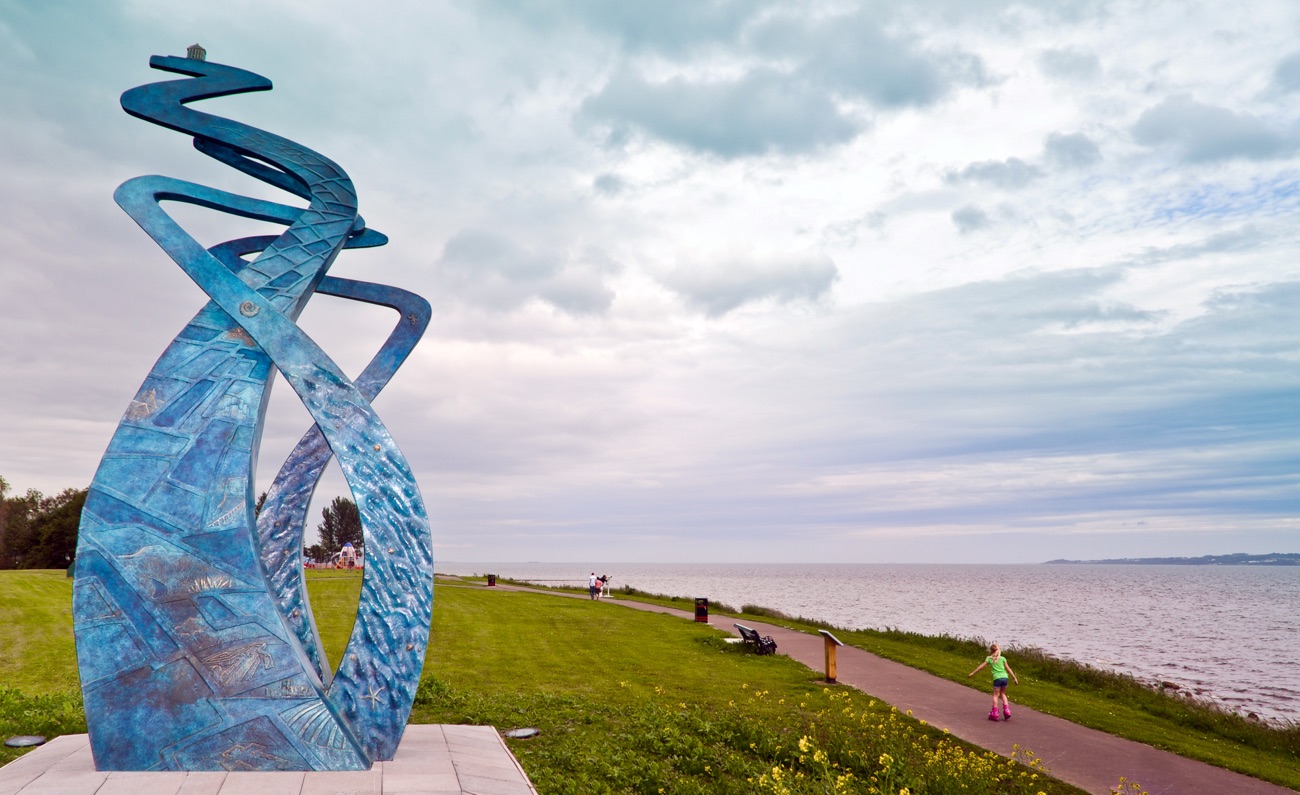 A tall blue mosaic sculpture on the grass beside the shore path at Jordanstown, with evening light over the lough