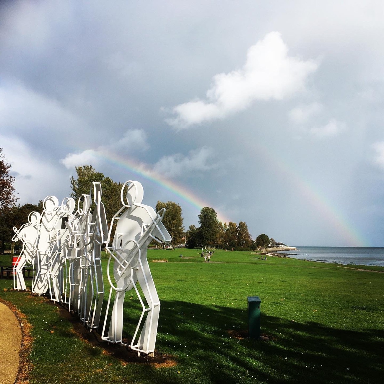 Cut-out figure sculptures on the grass with a rainbow arcing over Belfast Lough behind